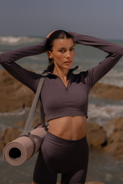 Woman in athletic wear holding a yoga mat on a rocky beach.