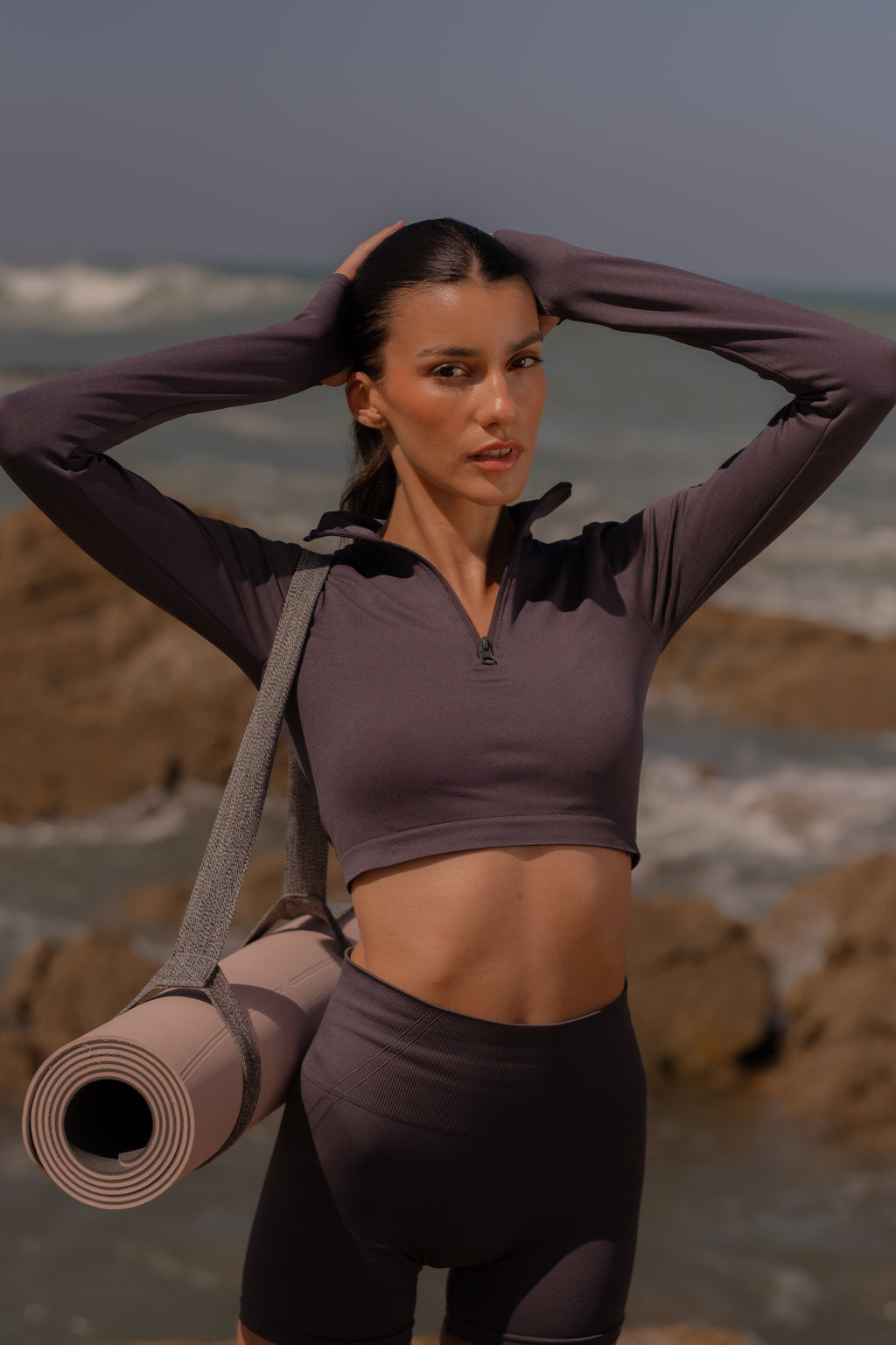Woman in athletic wear holding a yoga mat on a rocky beach.